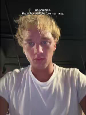 A selfie-style shot featuring a clean-shaven man with light blonde hair. He is wearing a white t-shirt. The background is out of focus but appears to be a dimly lit indoor setting. Text overlayed at the top describes a humorous take on marriage and relationships.