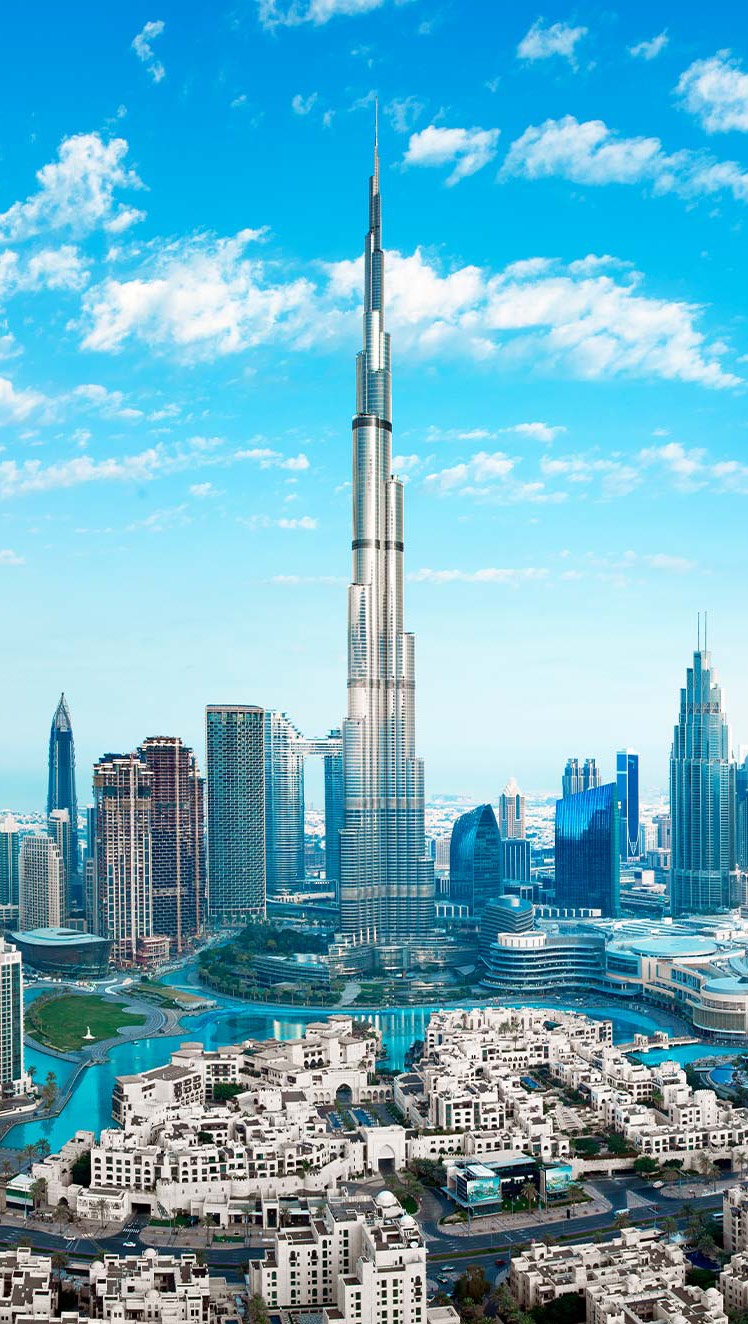 An aerial view showcases the iconic Burj Khalifa standing tall amidst a modern cityscape under a bright, cloud-filled blue sky. In the foreground, clusters of buildings with white and beige exteriors are spread out around a body of water. Above the buildings, a network of city structures extend towards the horizon with a rich assortment of glass-clad skyscrapers and other buildings. The overall vibe is bright and inviting, highlighting the architectural beauty and urban landscape of Dubai.