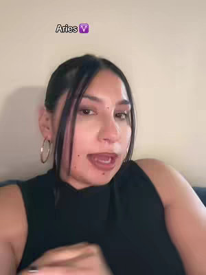 A selfie-style image featuring a woman with dark hair and hoop earrings looking at the camera, open-mouthed, in front of a plain wall. The text overlay at the top says 'Aries' with a purple TV symbol.
