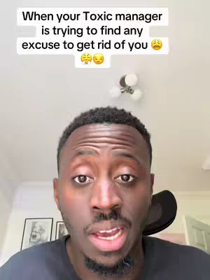 A man of color is centered in the frame, facing the viewer with an open mouth as if speaking. Above his head, a white rectangular box contains text in black, accompanied by a series of emoji faces. The background is a plain white wall with a light fixture and two framed pictures visible. The man is wearing a black shirt.