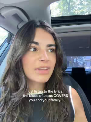 A woman with long brown hair is filmed from the chest up, sitting in a car. Text overlaid on the image reads a religious message.