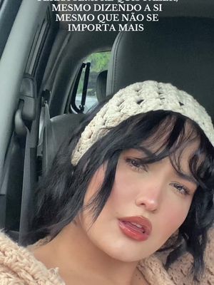 A close-up shot of a woman inside a car. The woman has dark hair, is wearing a cream-colored crocheted headband and a light colored top. The text overlay at the top in white text contains text in Portuguese.