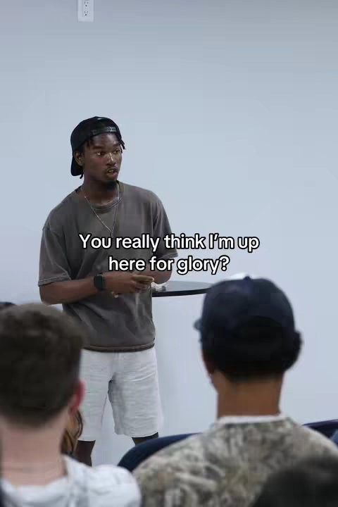 The image shows a man in a room, with a plain gray wall in the background. He has dark hair, wears a black cap, a grey t-shirt, and white shorts. Text overlays read 'You really think I'm up here for glory?' He appears to be addressing an audience, with several blurred individuals visible in the foreground.