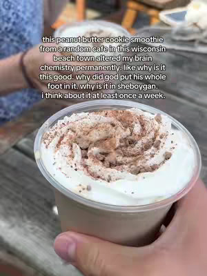 The cover image shows a close-up of a peanut butter cookie smoothie in a clear plastic cup. The drink is a light brown color and topped with whipped cream and a dusting of what appears to be cocoa powder or cinnamon. The cup is held in a hand, with a glimpse of the person's arm and a bracelet visible. In the background, out of focus, are hints of a wooden surface or table and other cups.