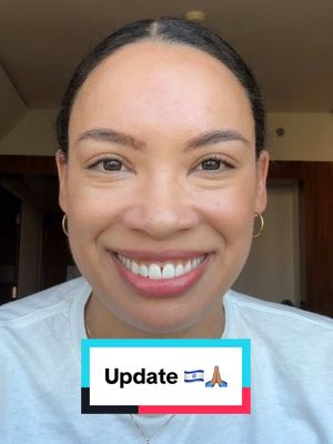 A smiling woman with light skin and dark hair is centered in the frame, framed by a medium shot. She wears a white t-shirt and gold hoop earrings. Her eyebrows are well-groomed, and her eyes are directed at the viewer. A colorful rectangular box with the word 'Update' and an Israeli flag image, along with praying hands, is overlaid at the bottom of the frame. The background is a blurry interior setting.