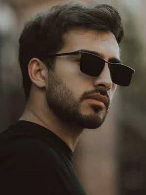 A close-up shot of a man wearing sunglasses. He gazes off to the side with a composed expression. The background is blurred, suggesting an outdoor setting.