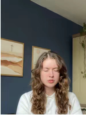 A young woman with long, curly brown hair is positioned in the center of the frame, with her eyes closed and lips slightly parted, appearing to be singing. She is wearing a white collared shirt. The background is a mixture of a darker blue wall, on which two pieces of framed art hang. To the right of the woman is a dark-colored wall with visible hints of green plants.