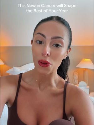 A woman with dark hair and red lipstick is centered in the frame, appearing to be filming a vlog. The background suggests a bedroom setting with a neutral color palette where two lamps are visible behind the woman. Text overlays at the top of the image.