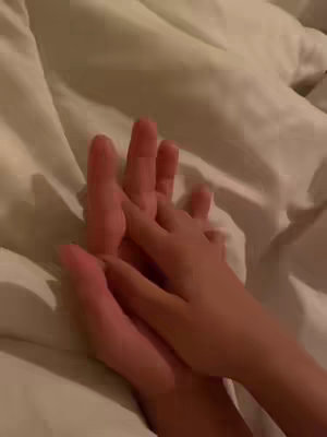 The image captures a close-up view of two hands intertwined, presumably resting on a bed. The larger hand, likely an adult's, cradles a smaller hand, possibly a child's, creating a tender and intimate composition. The hands are the focal point, and the blurred background suggests a soft, cozy setting. The lighting appears natural, casting gentle shadows and highlights that accentuate the textures of the hands and the bedding.