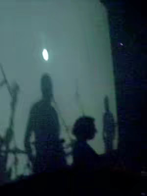 The image depicts a dim, teal-tinted scene, likely a live performance or concert. There are silhouetted figures suggesting band members and a possible audience. The lighting is subdued, creating a moody atmosphere. The composition is centered, with the silhouettes appearing against a lighter background that may be a screen or projection.