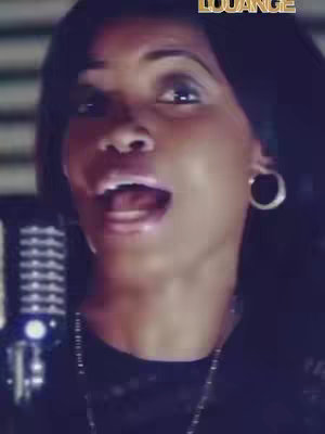 A close-up shot focuses on a woman's face. She has dark hair, hoop earrings, and a necklace. Her mouth is open as if singing. Behind her is a blurred, dark background, and there's text in the top right corner.