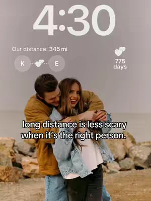 The image shows a couple embracing, with the woman laughing while the man hugs her from behind. The background has a blurred, neutral tone with hints of rocks. A time display and other informational overlays are positioned at the top.