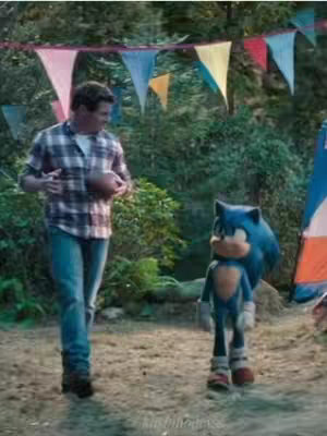 The image is a still from the Sonic the Hedgehog movie franchise. It presents a medium shot capturing a moment between the human character Tom Wachowski, seen on the left, and the animated character Sonic the Hedgehog, positioned on the right. Tom, a man, is outdoors in a setting decorated with festive triangular flags strung above in the background. He is dressed in a plaid shirt, jeans, and appears to be holding a football. Sonic, in his iconic blue hue, stands beside him, positioned close to Tom. The lighting suggests daylight conditions, and the color palette is natural, with blues and greens dominating, complemented by the red, yellow, and blue flags and the neutral tones of the setting. The composition balances the two figures, with the setting providing a cheerful backdrop.