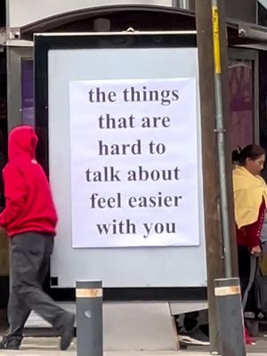 An urban scene featuring a large, white poster with black text displayed on a billboard. People are walking by in the background. The foreground shows a person wearing a red hoodie walking towards left.