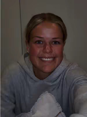 A selfie of a young woman with blonde hair, wearing a gray hoodie, and smiling at the camera. She has fair skin with a slightly reddish tint on her cheeks. The background has a plain, light-colored wall.