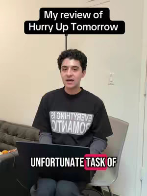 A man sits in front of a laptop, with text overlays at the top and bottom. He is wearing a black t-shirt with white text. The background is a plain white wall.