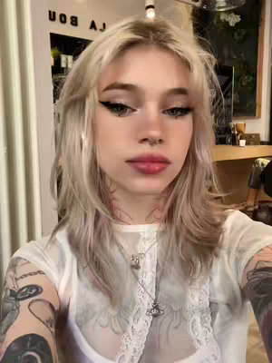 A selfie of a person after a haircut, smiling. The person has light hair styled in soft waves, and is wearing a white top. They've got noticeable eyeliner and a nose ring. The backdrop is slightly blurred, implying they are in a hair salon.