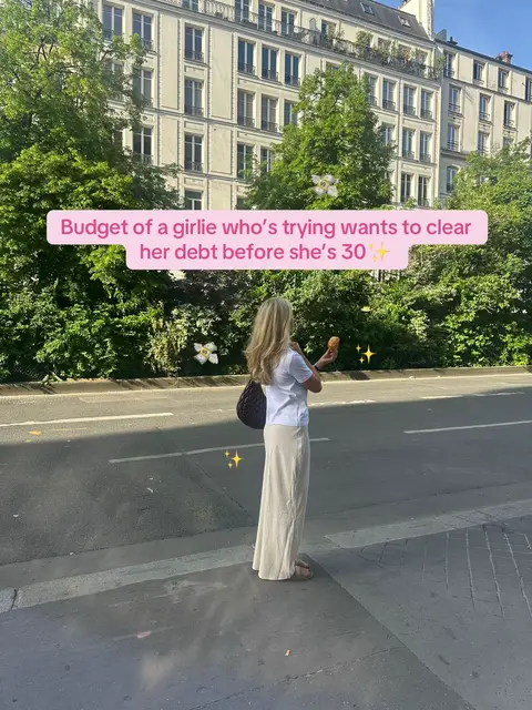 A woman stands on a city street, holding a small object. She's seen from the back, wearing a white shirt, a long beige skirt, and carrying a dark bag. The background shows a building, trees, and the street. A text overlay reads, 'Budget of a girlie who's trying wants to clear her debt before she's 30'.