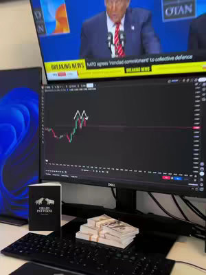 The image shows a desk setup related to finance and trading. A large computer monitor in the center displays a stock chart showing a candlestick pattern, flanked by a blue-tinged monitor on the left. A television at the top displays breaking news with Donald Trump. In the foreground, there are stacks of US dollar bills, a keyboard, and a notebook with a logo alongside the monitor displaying financial data.