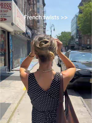A woman with blonde hair in an updo, seen from the back as she walks down a city street. She is wearing a black and white polka dot dress and carrying a brown purse over her shoulder. The street has buildings on either side and cars are parked along the street.