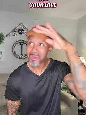 A man with a bald head wearing a black v-neck shirt and a tan beanie cap is gesturing upwards with his right hand in front of a domestic interior. The man is looking upwards with an expression of surprise.