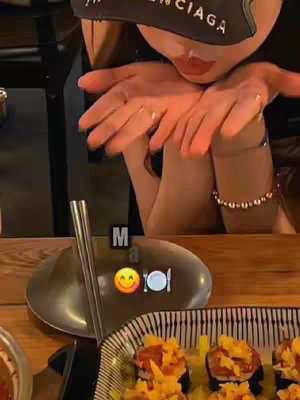 A person's face is partially visible, with the person wearing a black cap. The person appears to be blowing a kiss towards the camera, with sushi and a plate visible in the background.
