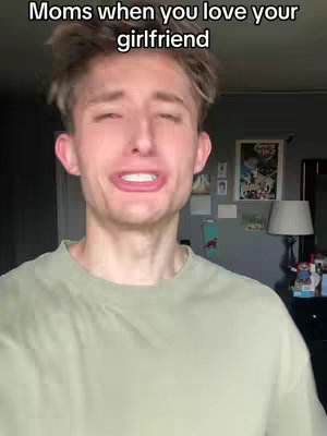 A young man with light hair is captured in a medium shot. He wears a light green t-shirt. His facial expression suggests distress or discomfort; his mouth is tightly pursed, and his eyes appear slightly watery. The background includes a glimpse of a room, with a gray wall adorned with framed images and a lamp.