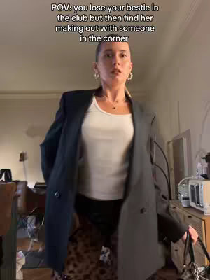 A well-lit shot features a woman in a casual outfit. The woman's face is in focus, displaying a look of slight bewilderment or surprise. The text 'POV: you lose your bestie in the club but then find her making out with someone in the corner' is overlaid at the top. The background is softly blurred, suggesting an interior setting.