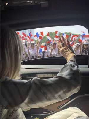 A woman in the foreground waving from the window of a car. Behind her, the view shows a crowd of people waving flags and cheering. The scene is brightly lit, suggesting a cheerful atmosphere.
