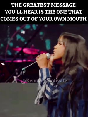 The image features a woman on a stage holding a microphone, with text overlayed at the top. The woman is center-aligned, wearing a dark blue blazer with a striped patterned shirt, and appears to be singing or speaking. Behind her are blurred stage lights and indistinct figures suggesting a large audience. The color palette is muted with darker tones.