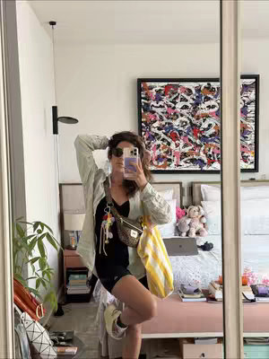 A woman is taking a selfie in a mirror. She is wearing sunglasses and a black top with shorts. She is holding a phone in front of her face. She is standing in a room with a bed in the background. There is also a painting and a window visible.
