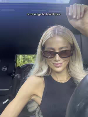 A woman with blonde hair and sunglasses sits in the driver's seat of a car, looking at the camera and smiling. Above her head is a text overlay against the dark interior of the vehicle, while outside, a green landscape is visible.