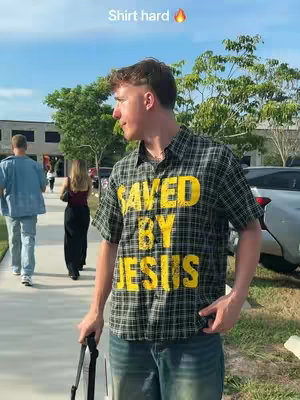 A young man wearing a plaid shirt with the words SAVED BY JESUS in large yellow letters stands on a sidewalk. He is looking to his left, with a serious expression. Other people are walking in the background. The sky is blue with a few clouds and some trees are in the background.