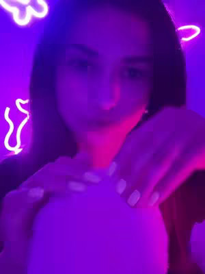 A close-up shot featuring a person's face, partially obscured by a large, light-colored sphere. The person is positioned centrally and appears to be engaging with the sphere using both hands. The scene is bathed in a purple and pink neon glow, with decorative neon shapes in the background.