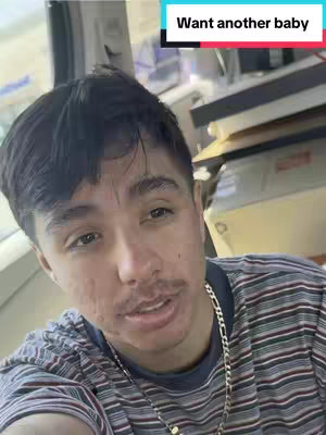 A selfie-style shot featuring a young man with dark hair and a mustache. He is wearing a striped shirt and a chain necklace. A text overlay reads "Want another baby" in a white font with a red and cyan background. The background appears to be an indoor setting, with some object behind him that look like paper.