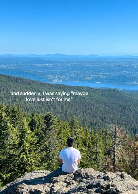 A scenic outdoor shot with a vast landscape of green hills and mountains under a clear blue sky. A person is seated on a rock, facing away from the camera, with their back to the viewer. The text overlayed says: "...and suddenly, I was saying "maybe love just isn't for me". This suggests a contemplative or reflective tone, possibly related to personal struggles shown in the video.