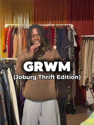 A man with dreadlocks stands in a clothing store, arms out, gesturing towards the camera. He wears a brown hoodie and light blue pants, with a clothing rack visible behind him and text overlays. The background features racks of clothing and decorative red curtains.