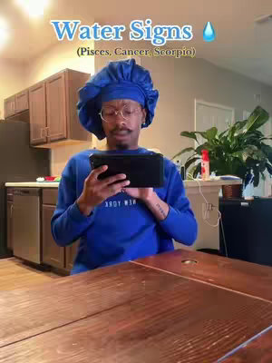 A person wearing blue head covering and attire is seated at a dark wooden table, holding a black tablet. Overhead text announces 'Water Signs,' accompanied by a water droplet icon and a list of astrological signs. The background includes a kitchen and a green plant.