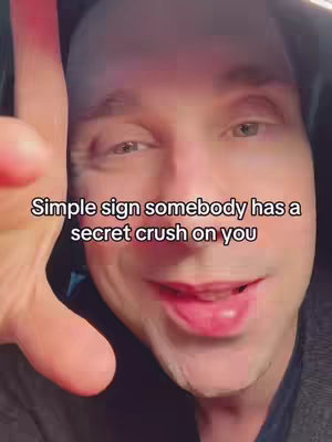 A close-up shot of a man's face filling most of the frame. The background is dark, and the man is looking towards the camera. Text at the bottom reads, 'Simple sign somebody has a secret crush on you.' His hand partially obscures the upper left.