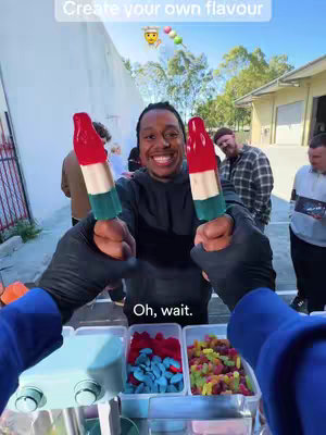 A first-person view of a man holding two large ice pops seemingly of different colors, with a variety of candies in containers in front of him. In the background are several people, and text overlays the top of the cover image.