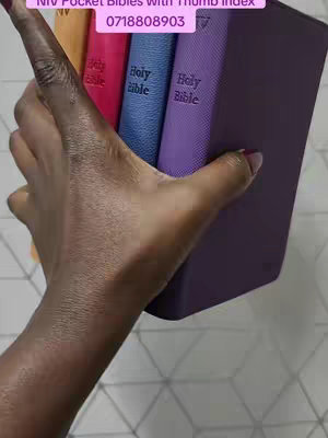A hand holds a stack of pocket Bibles in various colors, against a light-colored diamond-patterned background. The hand is dark-skinned, with painted nails, and the bibles include purple, blue, pink, and yellow spines. Text overlays at the top read details of the product, with a phone number.