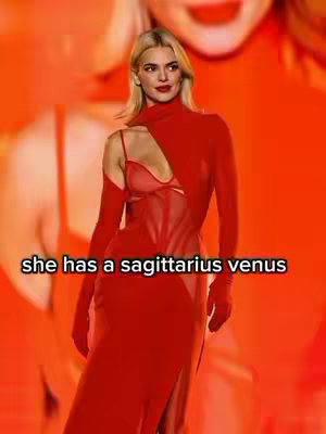 A woman in a red dress is standing in front of a red background. The dress features a high neck, a cutout on one shoulder, and a long, flowing skirt. The background appears to be a blurred image of a similar person or a repeated design.