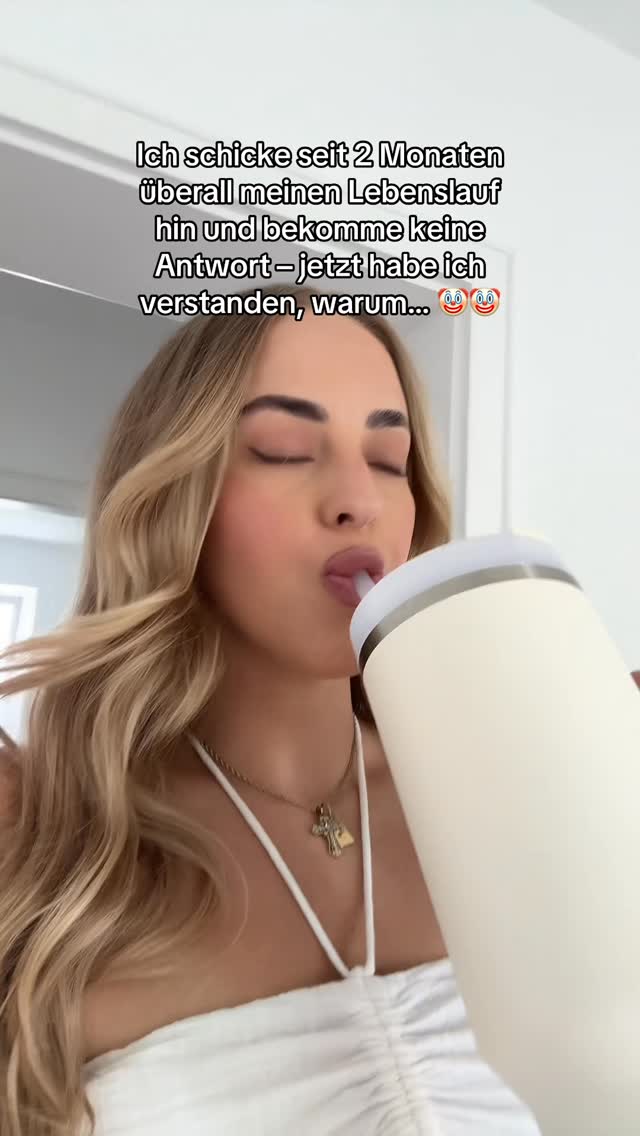 A woman with blonde hair is shown in a close-up, slightly above the shoulders view, sipping from a white insulated tumbler. The upper half of her face is visible, showing closed eyes and pursed lips as she drinks through a straw. She is wearing a white top and a gold necklace with a small pendant. The background appears to be a bright, softly lit interior.