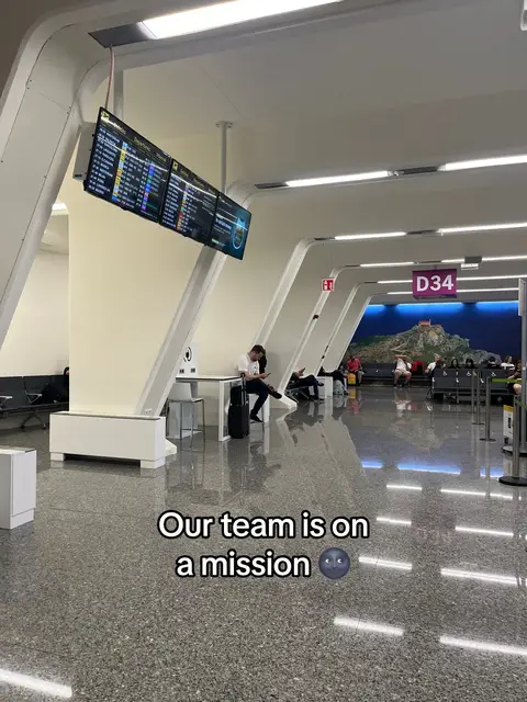 The image shows an airport terminal with a gray tiled floor. The ceiling consists of white, angular structures with inset lighting. A large information board hangs from the ceiling, displaying flight information. In the foreground, there's text overlaid, and a person is sitting near the boarding area. The overall feel is modern and clean.