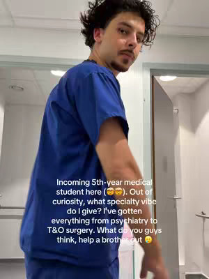 A young man in blue medical scrubs stands indoors, slightly angled to the camera. He has dark, curly hair, a mustache, and a focused expression. The background includes a hallway and a doorway.