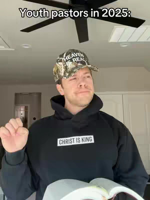 A man in a black hoodie and a baseball cap is holding a book in front of a ceiling fan, there is text across the image.