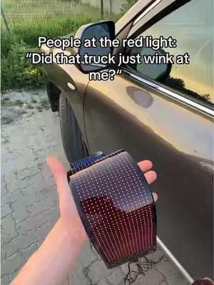 A person's hand holds up a car tail light modification. The tail light is off-white and features a matrix array of lights and a red-tinted housing. The background includes a car door and the side mirror. Text overlay reads "People at the red light: 'Did that truck just wink at me?'"