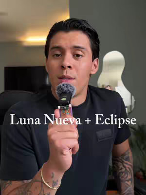 A man with short, dark hair is holding a microphone, with the text "Luna Nueva + Eclipse" overlaid at the bottom of the frame. He is wearing a black t-shirt and a gold bracelet, and has tattoos on his forearm.