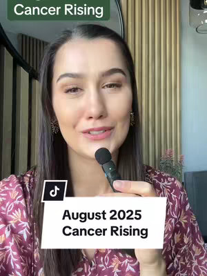 A woman with long dark hair and makeup is holding a microphone and speaking to the camera. The background includes a wooden wall and a decorative mirror. Overlay text reads "August 2025 Cancer Rising" in a white rectangular box with a TikTok logo.