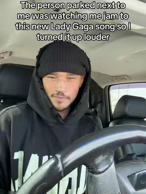 A man in a black hoodie and beanie sits in a car holding the steering wheel. Text overlays read the person parked next to me was watching me jam to this new Lady Gaga song so I turned it up louder.